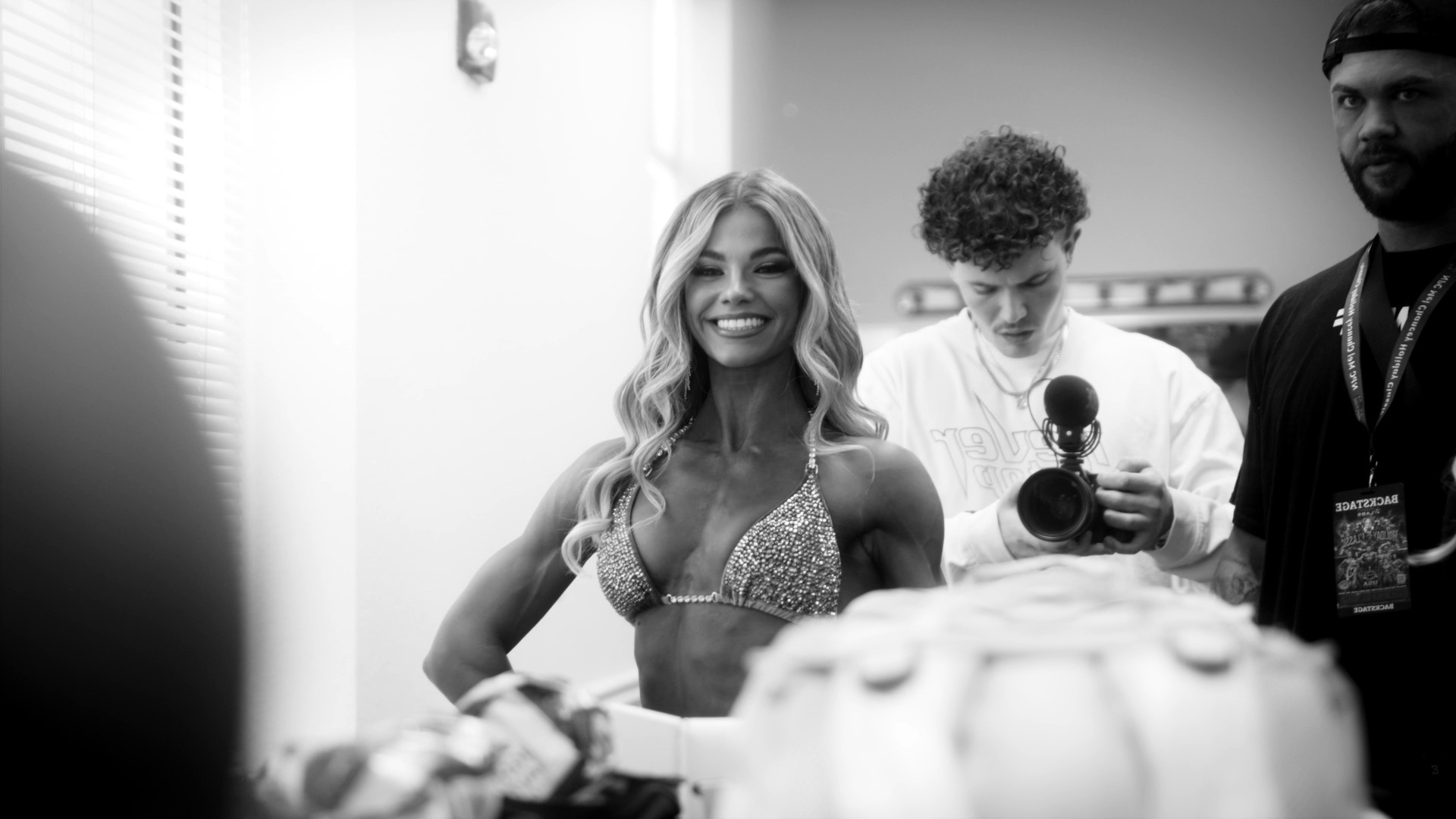 Backstage at a bodybuilding show: a competitor smiling for the camera while Grayson films behind her